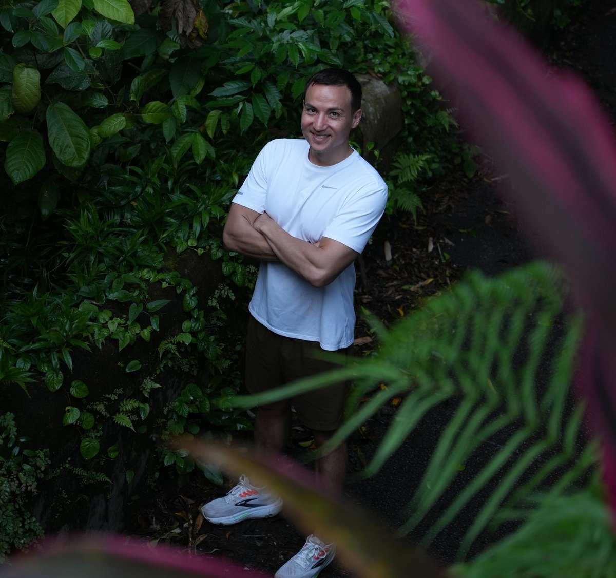 Chris standing among green foliage, smiling at camera, wearing white t-shirt